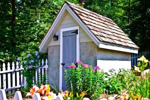 A small garden she shed with flowers blooming around it and a white picket fence.