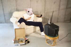 Man with pink pigs head in basement watching TV eating pizza and drinking beer to how man cave