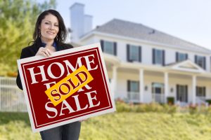 Smiling Hispanic Woman Holding Sold Home For Sale Sign In Front of Beautiful House to show the pitfalls of letting a home slip by
