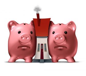Housing crunch and home crisis concept as two piggy banks putting the squeeze on a family house as an economic symbol of feeling financial pressure and finance stress from realestate mortgage prices and renovation expenses.